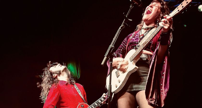 Tom Shemer kneels before his Lee-la Baum as the Damn Truth perform live onstage. He wears a red shirt and plays a Les Paul while Baum plays a retro single-cut electric and screams into the mic.