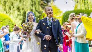 A newly-wed couple walking down the aisle hand-in-hand smiling whilst being showered by colourful rose petal confetti