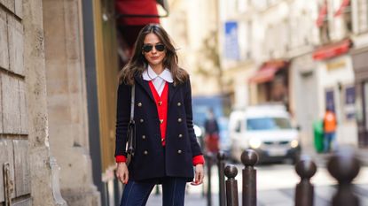 Therese Hellström wears Ray-Ban sunglasses, a white shirt with Peter Pan collar, a navy dark blue oversized military wool jacket with golden buttons, a red wool cardigan with golden heart-shaped buttons, a brown leather Hermes bag, blue denim jeans, beige quilted shoes, on March 19, 2021 in Paris, France