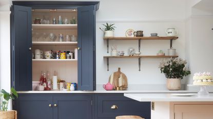 Should a kitchen island match the cabinets? Here is a kitchen with dark blue kitchen cabinets, with one opening up to display glassses and mugs, two wooden shelves, and a pink kitchen island with a white countertop