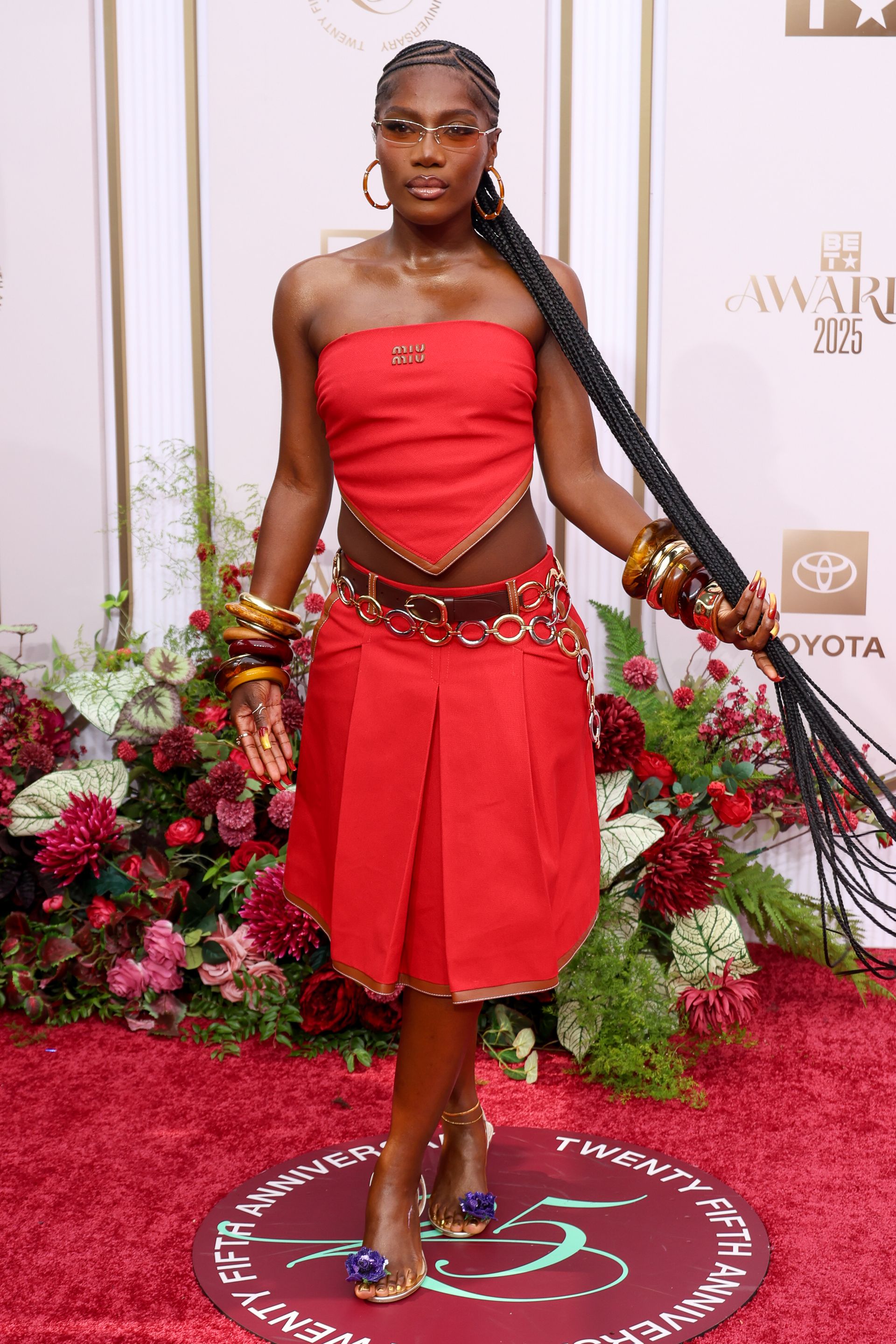 Woman wearing a red scarf top and red pleated skirt, floral heels, resin stacked bracelets.