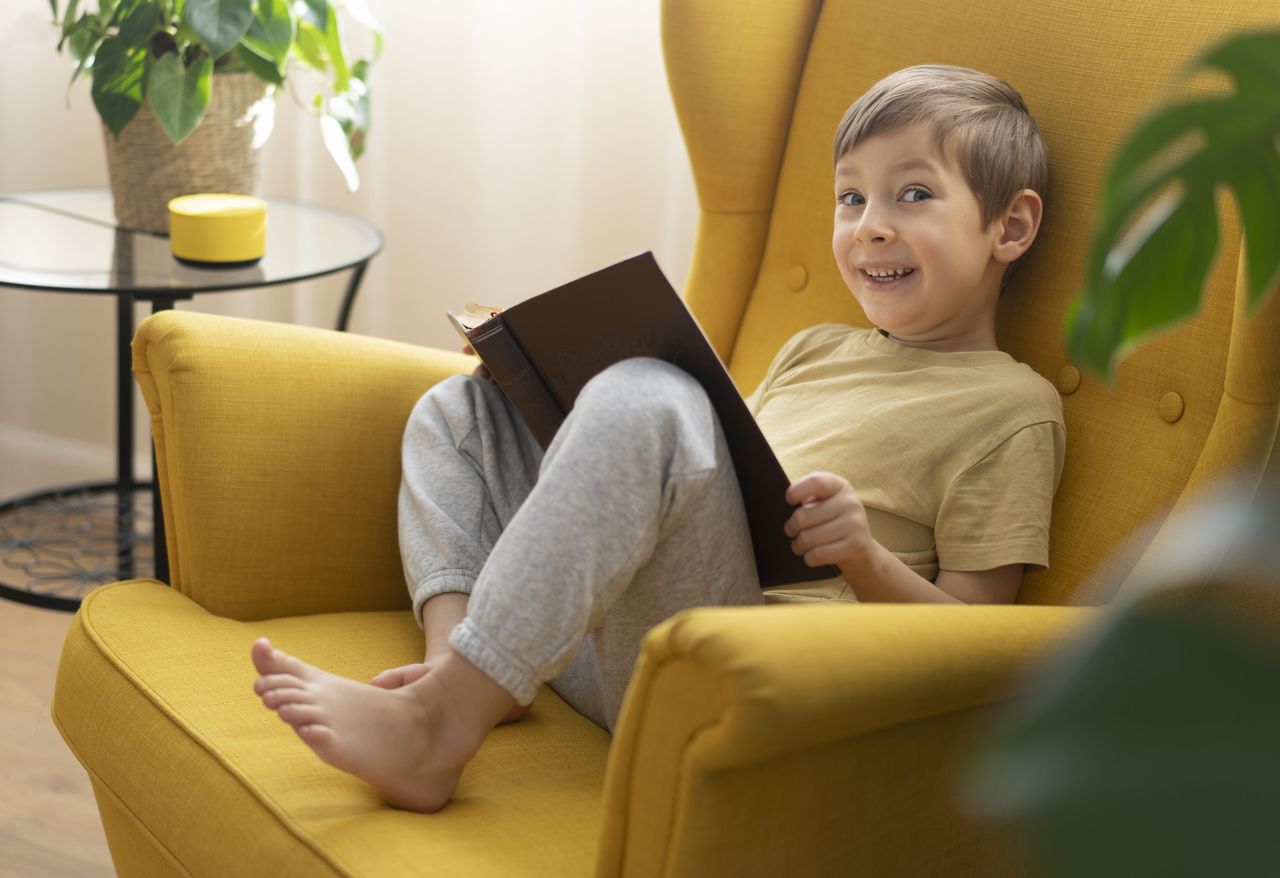 A little boy sits in a chair with the book and communicates with a smart speaker - stock photo