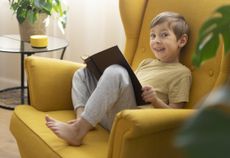 A little boy sits in a chair with the book and communicates with a smart speaker - stock photo