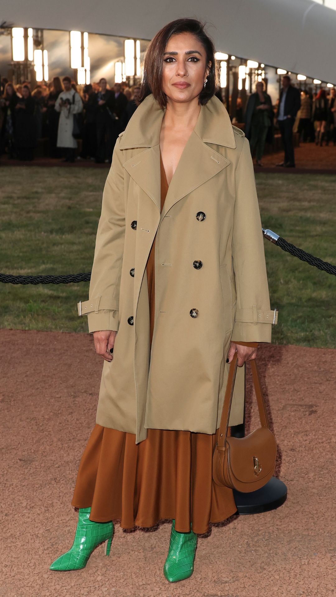 Anita Rani wearing a Burberry trench coat over a copper Victoria Beckham dress, carrying a tan Burberry bag and wearing emerald green faux crocodile skin boots