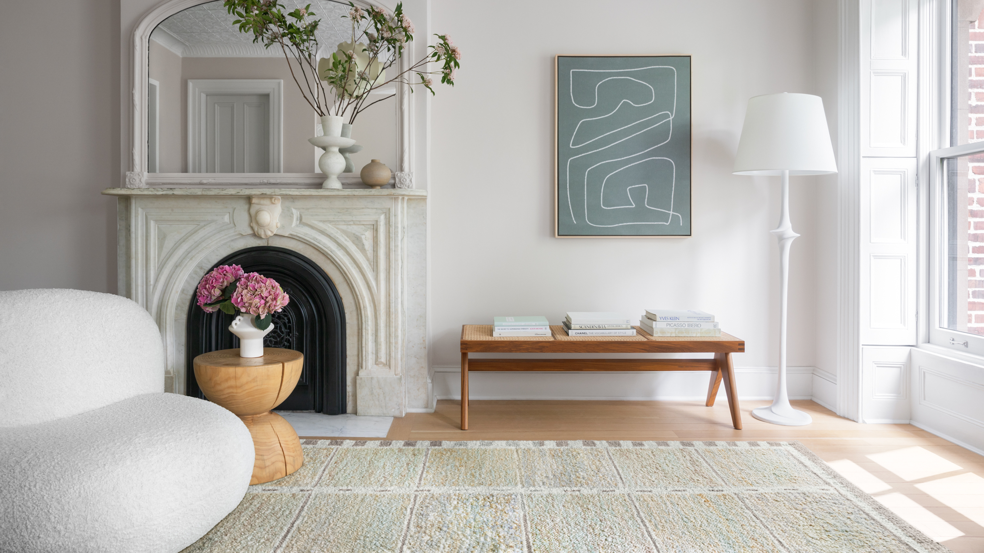 Living room with cream colored textured area rug, curved armchair, wooden side table and bench, fireplace with mirror above and floor lamp