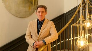 Rob Brydon standing on a sweeping golden staircase with a cream jacket and navy shirt on 