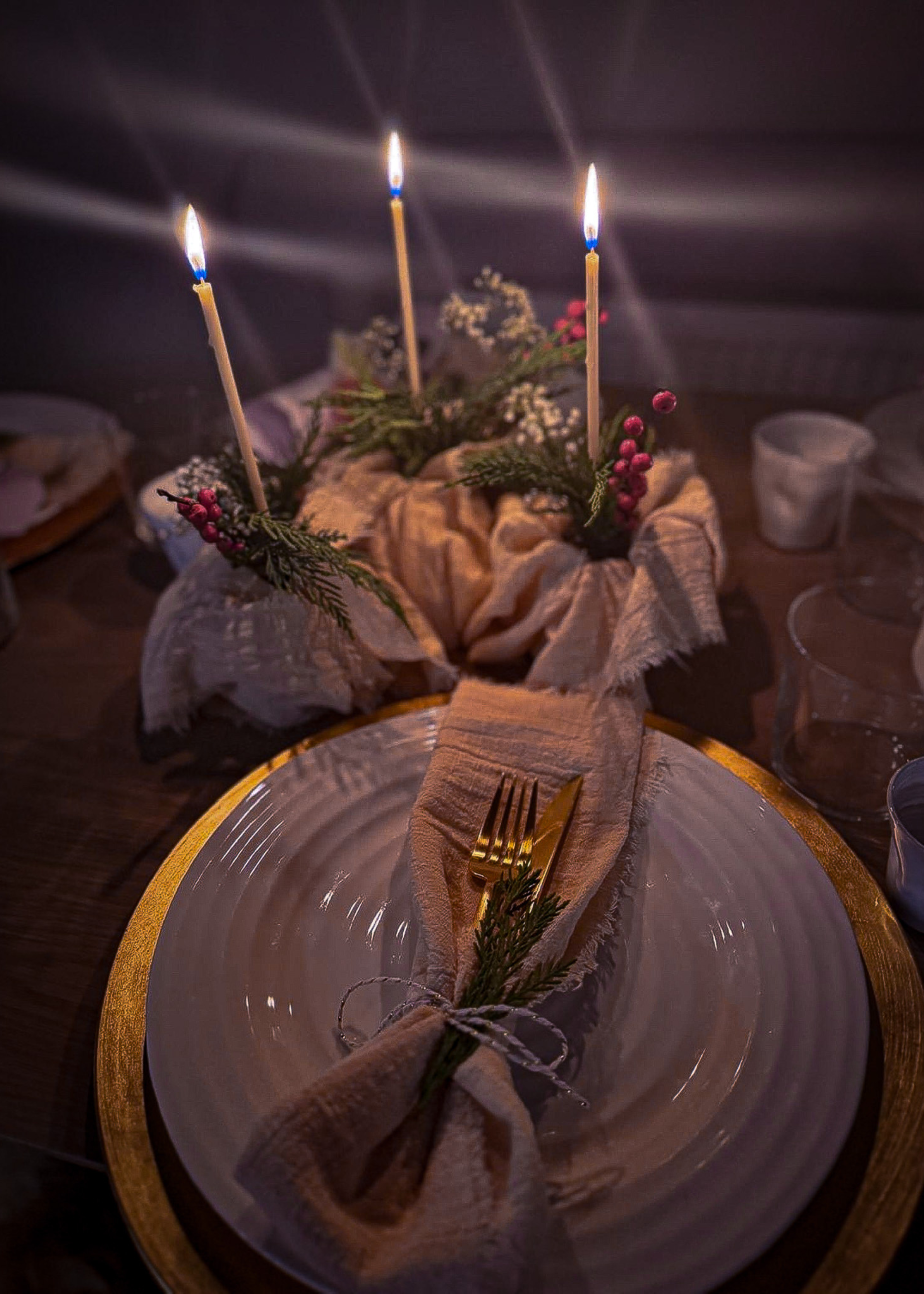 A christmas place setting with a napkin and gold cutlery wrapped in string and a trip of thin candles wrapped in a muslin napkin with foliage as a table centerpiece