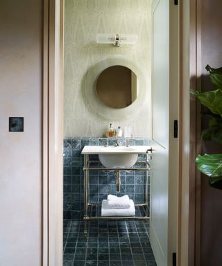 Bathroom with round mirror over Victorian washstand, Paisley patterned light green wallpaper an grey wall and floor tiles.