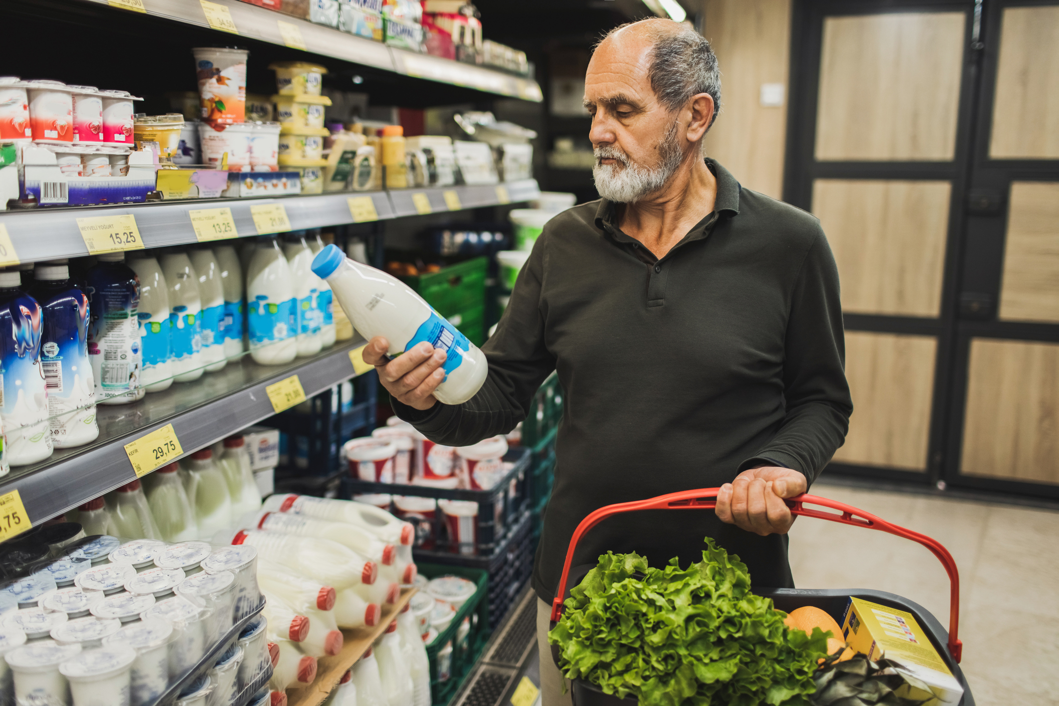 Older man buying milk and looking at the price. Holding basket with fresh lettuce and vegetables. Living on retirement.