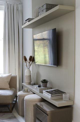 a neutral living room with a tv on the wall and long shelves above and below, with cream footstools tucked under the bottom shelf