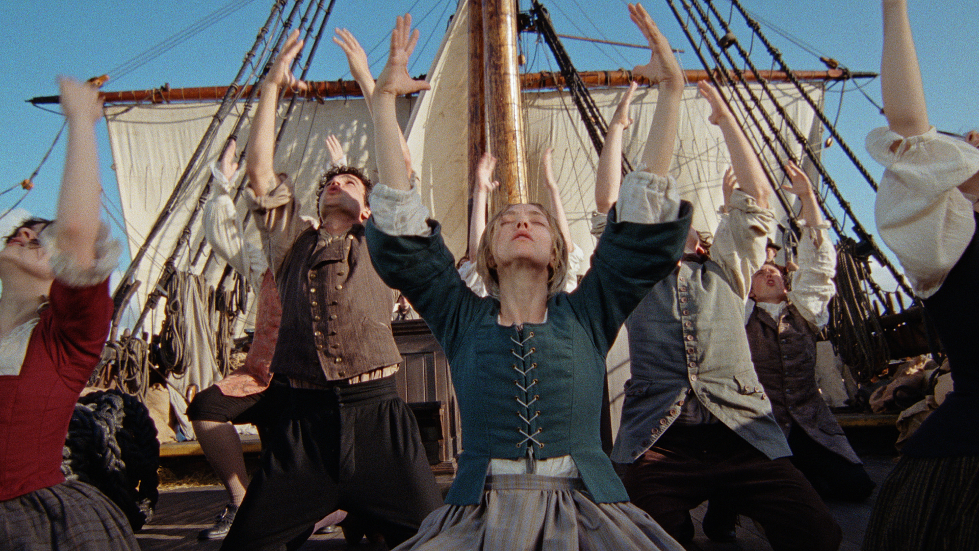 A group of people (center: Amanda Seyfried as Ann Lee) perch on their knees and raise their heads and hands to the sky. They wear 18th century clothing as they dance on the deck of a ship with a beige sail rising behind them, in THE TESTAMENT OF ANN LEE.