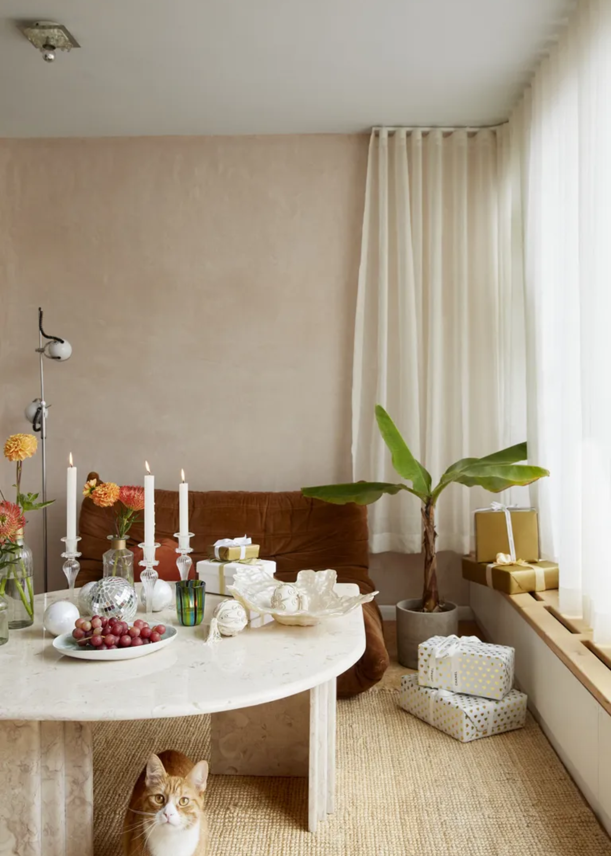A living room with a brown Togo, a potted tree, wrapped presents, a marble round table with tapers, a plate of grapes and a cat below