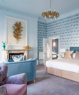 A bedroom with a white ceiling with a gold chandelier, blue and white patterned wallpaper, a white and gold leaf artwork, a teal blue velvet bed with beige pillows and white bedding, and a purple velvet floor