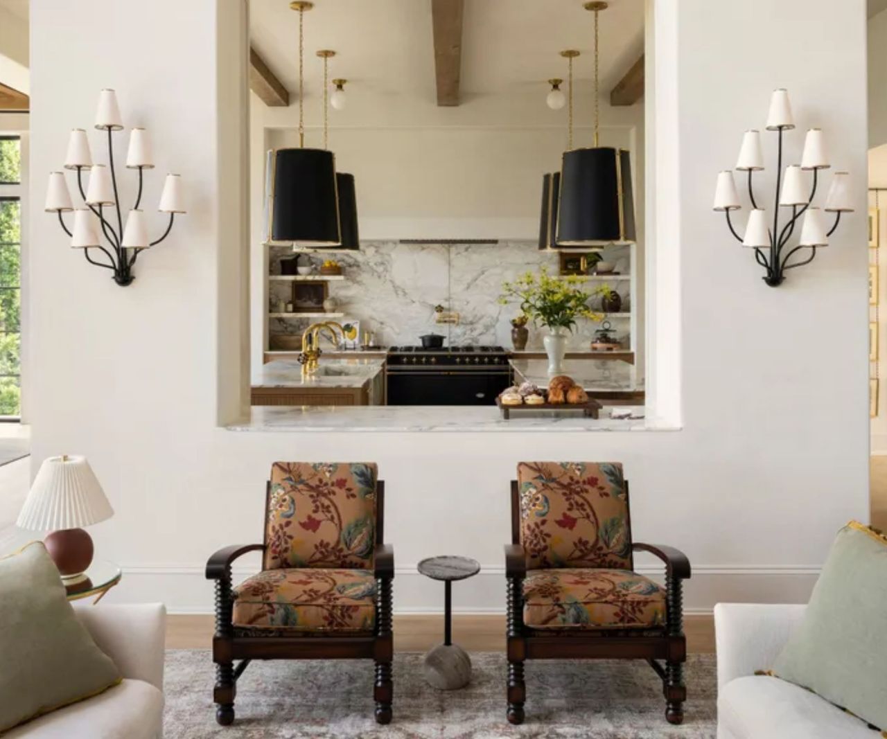 A bright living room with two white sofas facing a stone fireplace. A large tree sits in the corner, and shelves with pottery line the fireplace. A round gold chandelier hangs above, and tall black-framed windows let in natural light. A coffee table with books and flowers sits in the center.