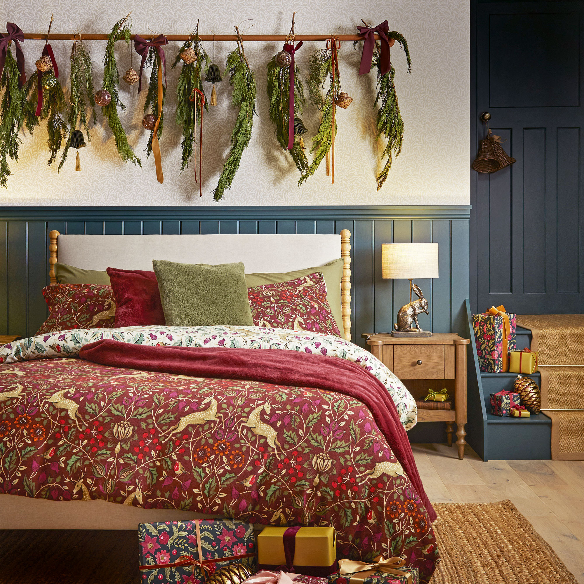 White and blue panelled bedroom with an oak bed with red arts and craft print bedding. A rail decked with pine branches and ribbons hangs over the bed.