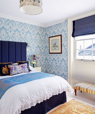 Bedroom decorated in pale blue and white, blue and white floral wallpaper, upholstered blue headboard, white bedding with blue throw, multi-colored scatter cushions, blue blinds, framed artwork on wall