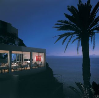 A large, modern hotel room is lit up at night to a backdrop of palms and beach