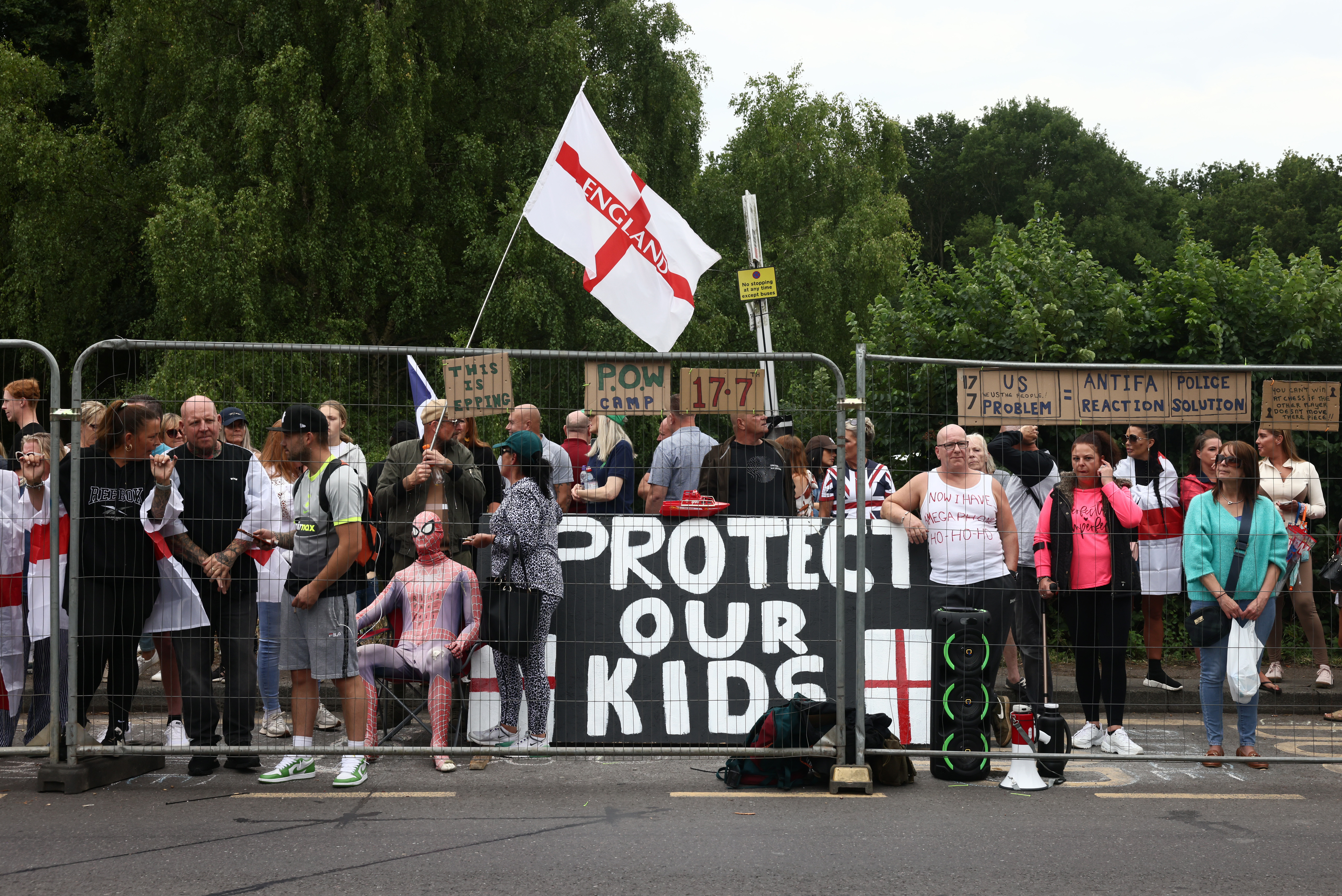 EPPING, ENGLAND - JULY 27: Protesters stand behind a fence as they gather near the Bell Hotel during a protest against the use of the hotel as migrant accommodation on July 27, 2025 in Epping, England. Anti-migrant protesters, as well as counter-protesters, have organised rallies in Epping near the Bell Hotel, which is being used as migrant accommodation. The far-right activist Tommy Robinson told his social media followers earlier this week that he would also join the demonstrations, although he did not attend. A series of protests have taken place here following the arrest of an asylum seeker on suspicion of alleged sexual assaults in the town.