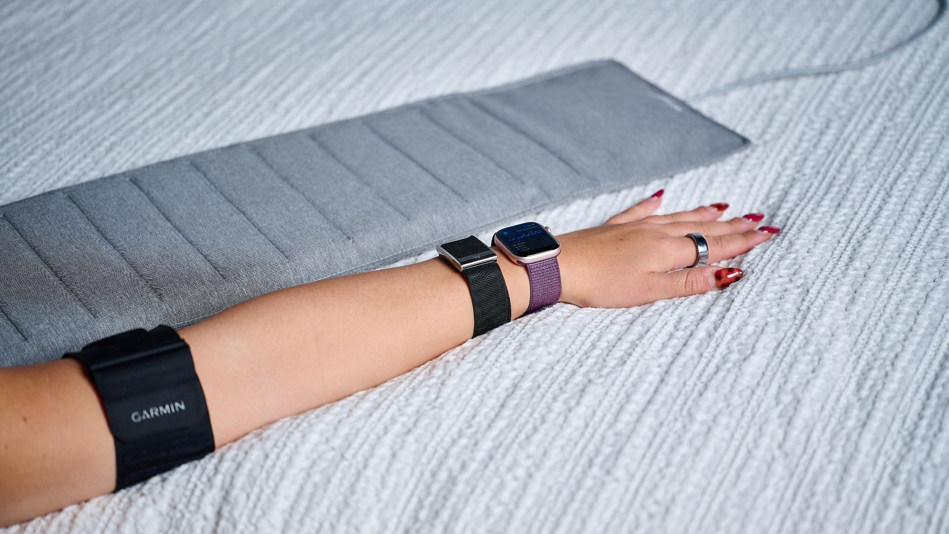 Our sleep tech tester wearing the Oura Ring 4, Apple Watch 10, Whoop MG and Garmin Index Sleep Monitor, holding arm out next to Withings Sleep Analyzer mat on white bed sheets