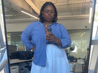 Chichi wears blue striped button down and white skirt while taking a mirror selfie in theBest Knockoff Luxury Clothing
 office.