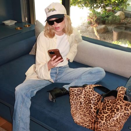 model Elsa Hosk sitting on a sofa looking at her phone while wearing an oversize baseball cap, black sunglasses, a pale yellow button-down shirt, white T-shirt, jeans, leopard-print bag, and black heeled boots