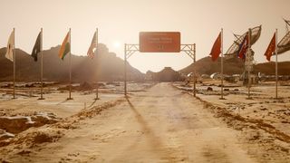 Still from the T.V. show For All Mankind (season 4, episode 1). Wide shot of a red desert landscape to represent the planet Mars. A dirt road leads up to a large sign which reads "Happy Valley." On either side there are several flag poles with different flags and to the right there are two large satellite dishes. There are some mountains in the distance.