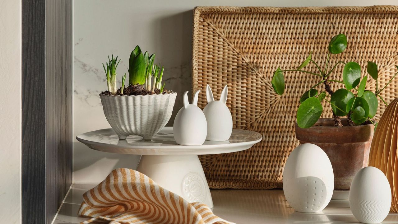 A white kitchen counter with Easter decorations on a cake tray, beside some freshly sprouting plants and flowers. 