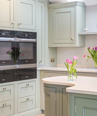 A kitchen with pale green kitchen cabinets with two built-in ovens and a curved kitchen island with matching cabinets