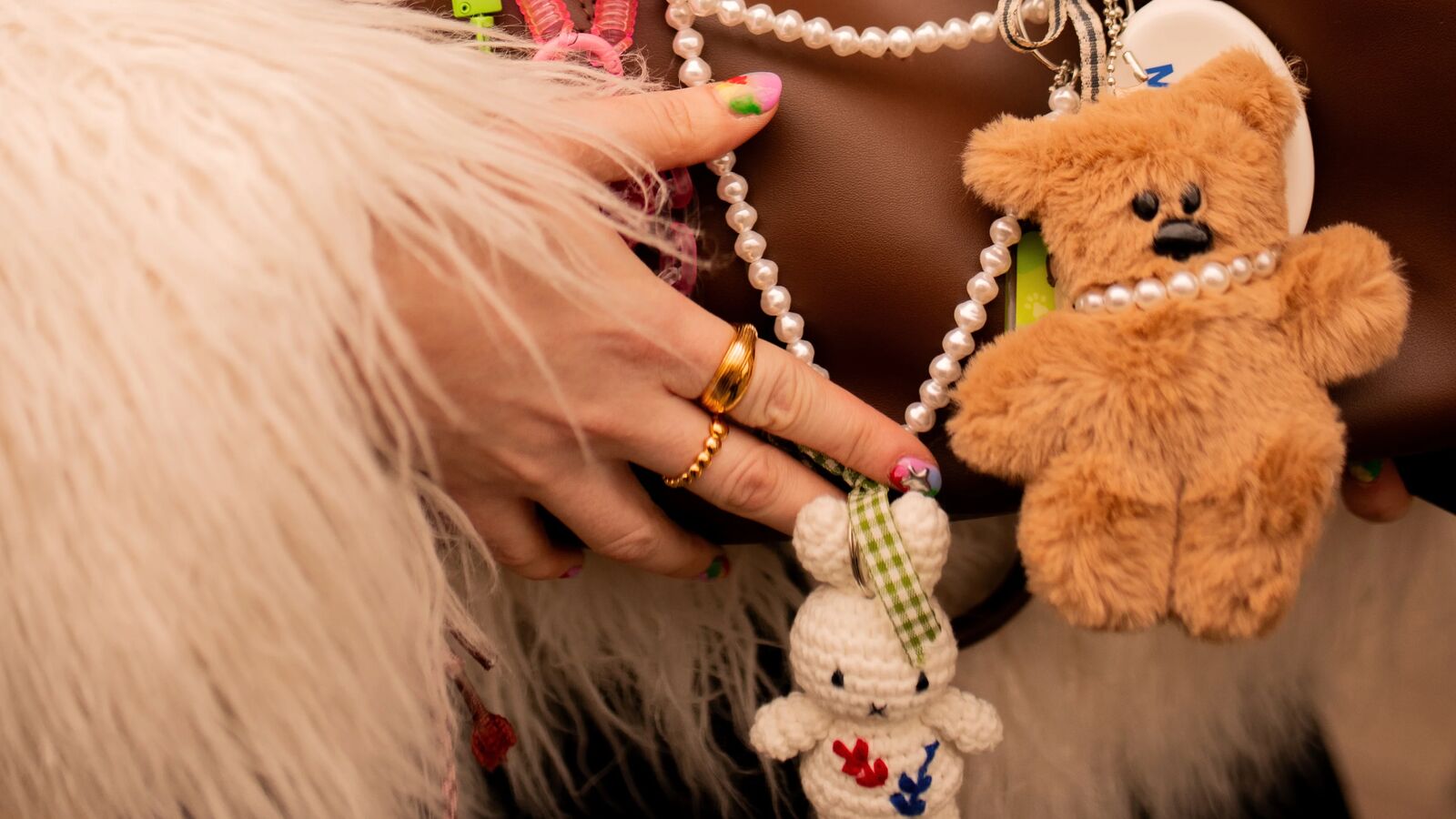 A woman&#039;s hand holding a brown handbag with various charms and decorations, including a teddy bear charm and a bunny charm with some faux pearls. 
