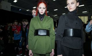 A red haired girl looks directly at the camera wearing a green jacket, in a queue of models