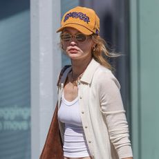 Gigi Hadid wearing a mustard yellow baseball cap, a collared cream cardigan over a white tank top, and cream drawstring chino pants with flip-flops in NYC