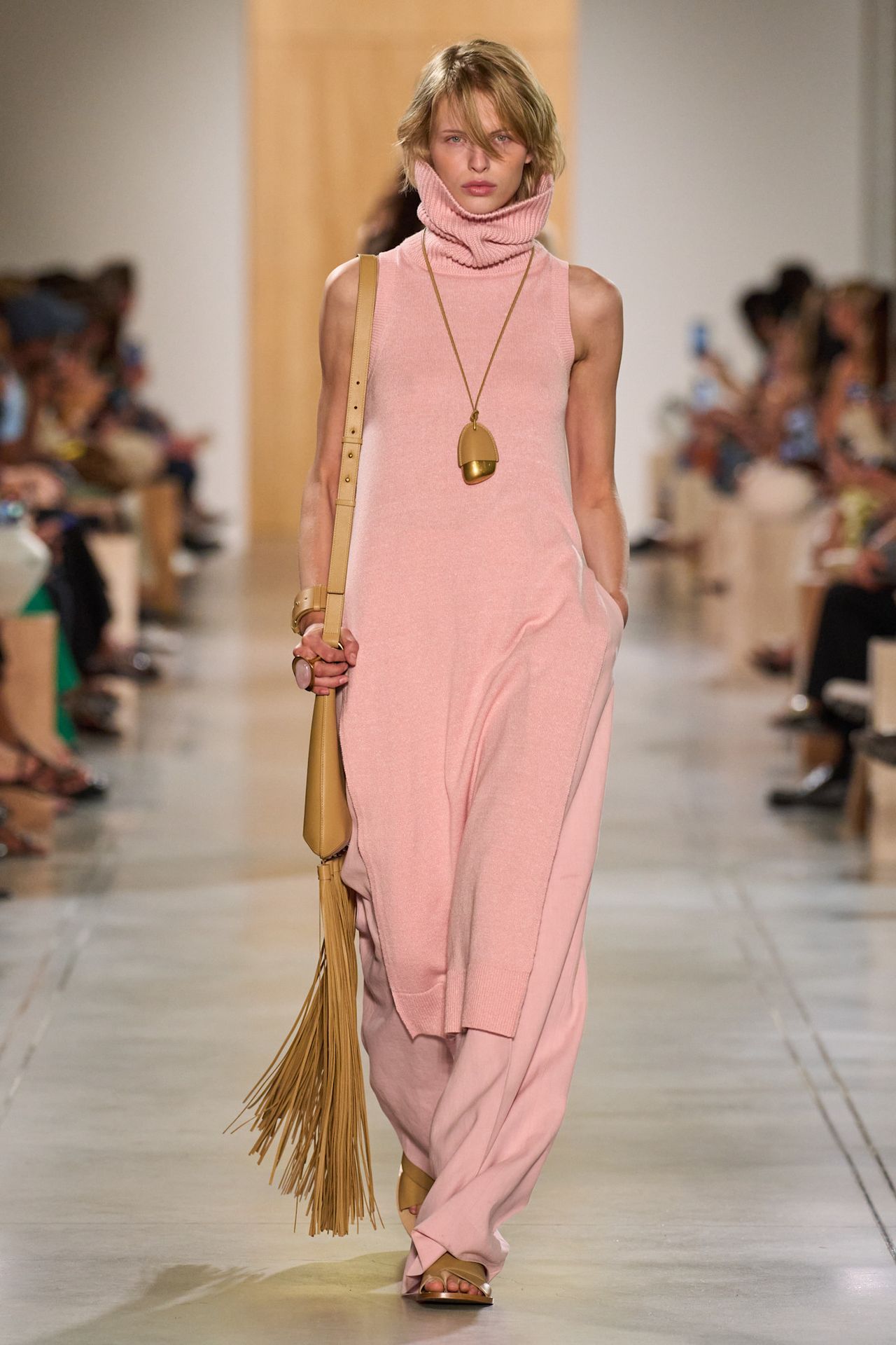 A Michael Kors model wearing a pink sleeveless turtleneck sweaterdress layered over matching wide-leg linen trousers, light tan slide sandals, a light tan fringe shoulder bag, a tan and gold long cord necklace, and a light pink gemstone ring.
