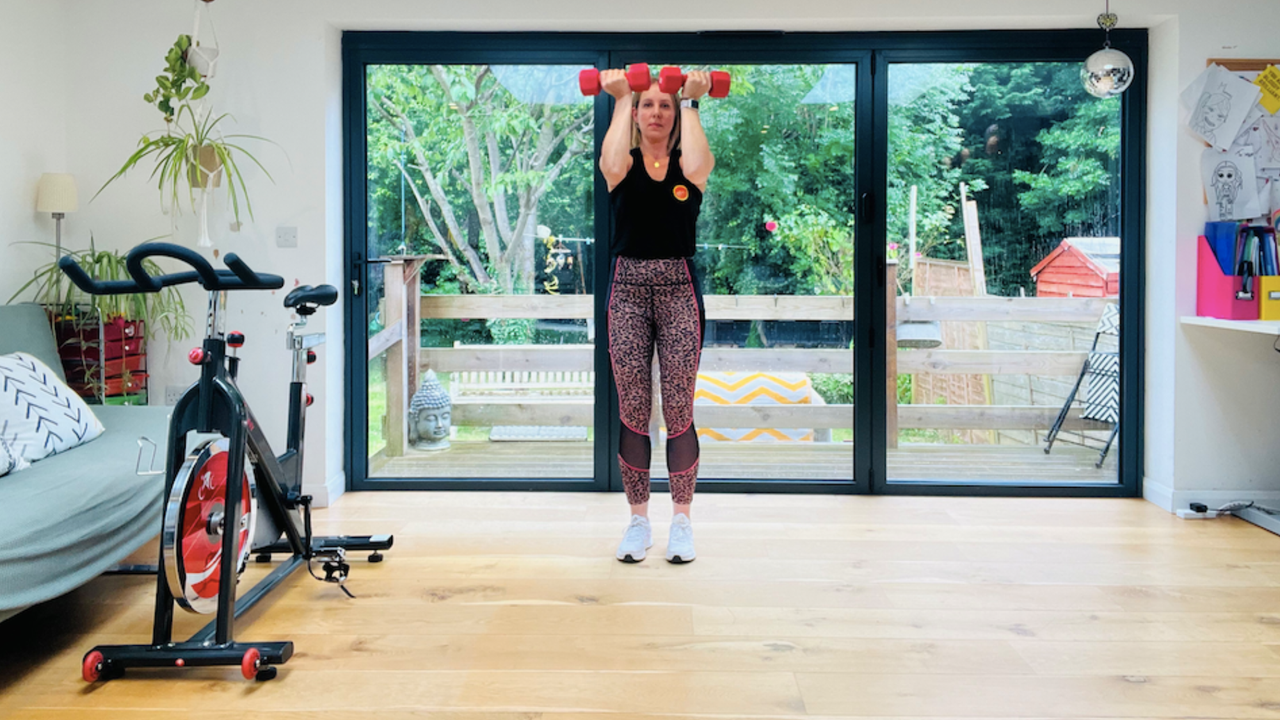 Personal trainer Maddy Biddulph performs the first part of an Arnold press in her well-lit living room. She wears a black vest, leopard-print leggings and white sneakers. She is standing and holding a pair of light dumbbells, with her elbows bent and the dumbbells close to her head. Behind her is a window showing a leafy garden and next to her is an exercise bike.