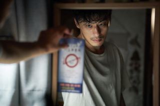 a man (Koo Kyo-hwan as Seol Kang-woo) is reflected in a mirror has he holds up a brochure, in 'parasyte the grey'