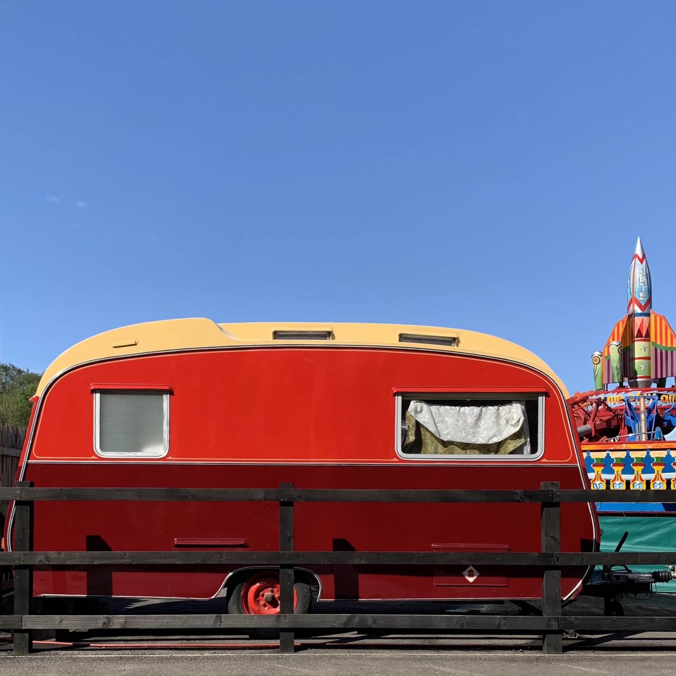 A red and cream vintage caravan parked behind a wooden fence, with a fairground rocket ride visible in the background.