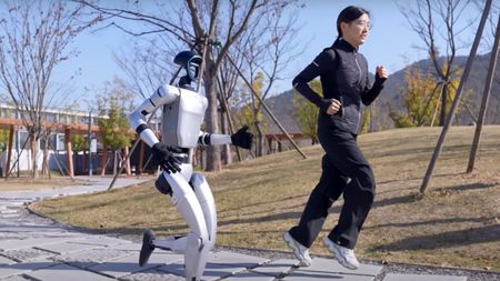 Bipedal robot running alongside a woman on a park path.