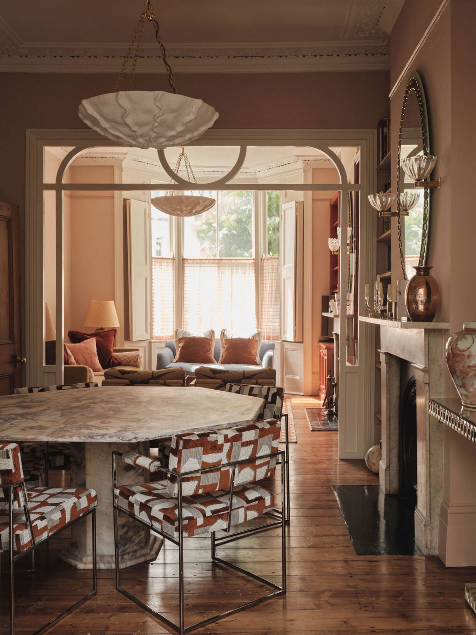 modern dining room with round marble table, patterned chairs, ornate fireplace with mirror above and wall sconces, and modern pendant light above, with door through to living room