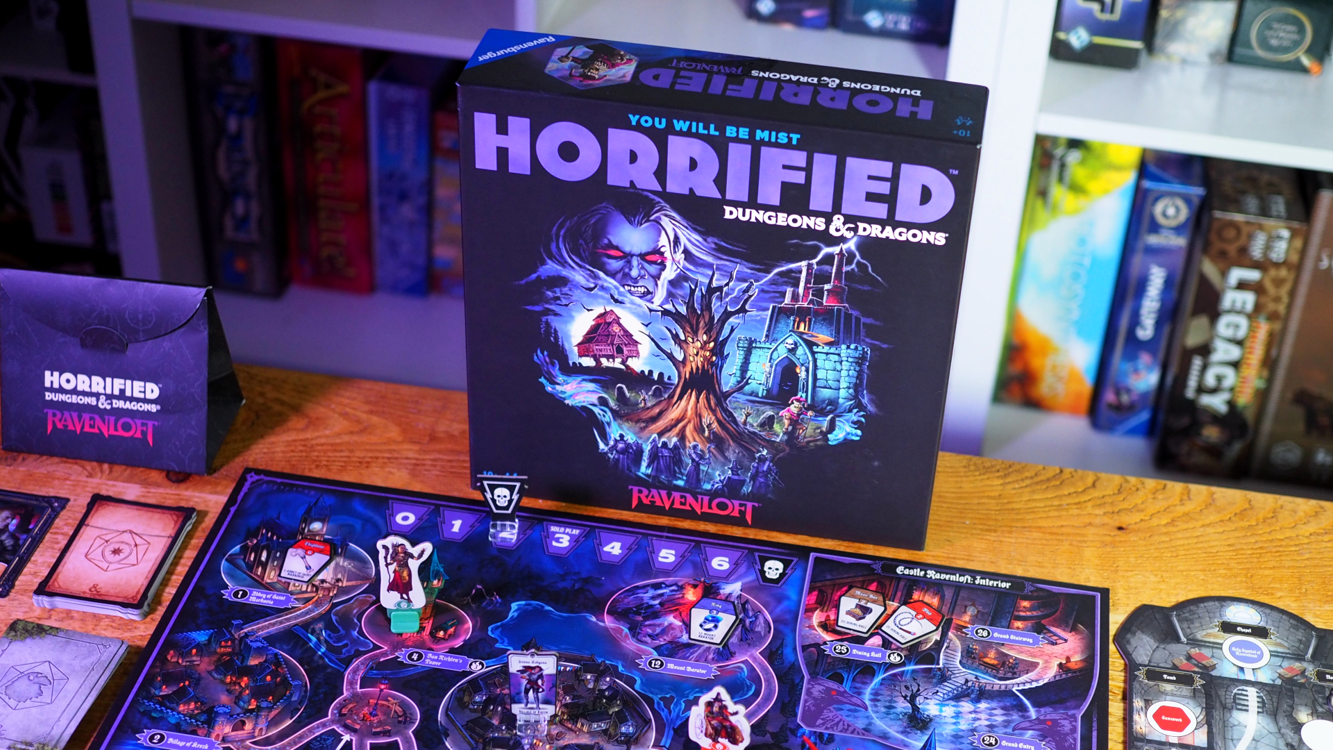 Horrified Ravenloft on a wooden table behind the set up board, with a shelf of board games slightly blurred in the background