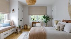 White bedroom with reading nook and vanity area, each of which is by well-lit and wide windows. There are linen and wooden accents, giving the room a bright but cozy feel