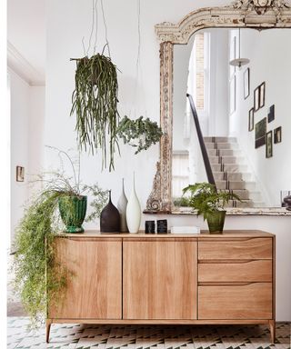 White hallway with mono graphic floor tiles, honeyed wood sideboard, oversized wall mirror, and variety of fresh plants and greenery at different levels.