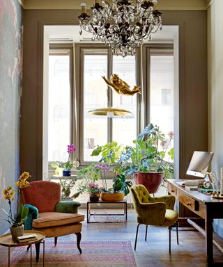 study with chandelier, oversized floral wallpaper, brass pendant lamp, velvet armchair, velvet office chair, wooden desk, patterned rug and plants