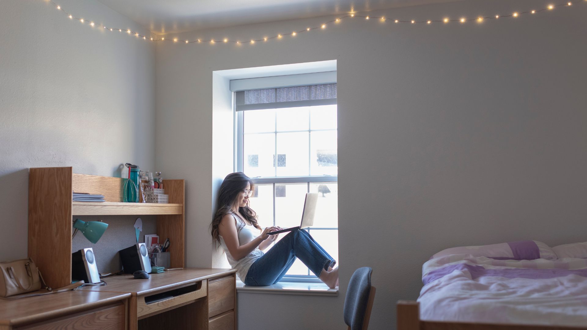 A female student sits on a window sill studying with a laptop in a college dorm room.