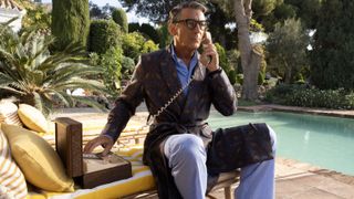 Jason Isaacs as Cary Grant in a patterned dressing gown sits on a yellow sunbed on the phone by the pool in Archie.