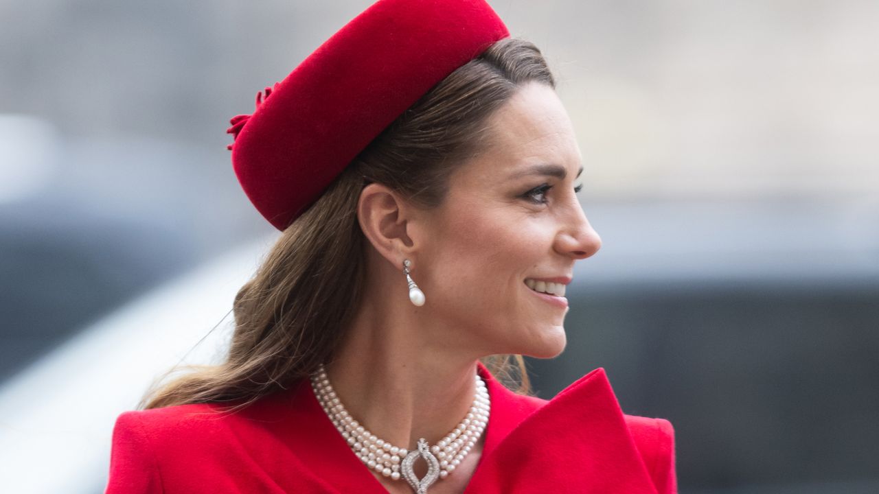 Kate Middleton wearing a red hat and coat with pearl necklace turning to the right and smiling