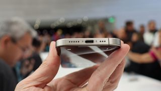 En hand håller upp en iPhone 15 Pro Max vid ett lanseringsevenemang och visar upp telefonens undersida och laddningsport.
