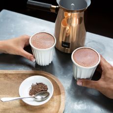 Hotel Chocolate Velvetiser on table and two people holding mugs with hot chocolate in