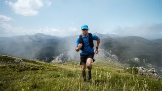 Mature sportsman trail running in the mountains on meadow against blue sky