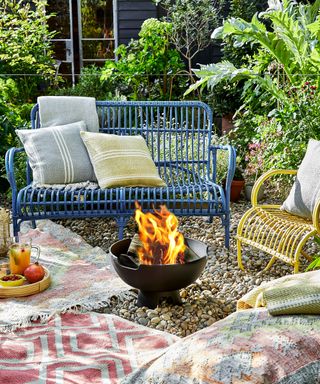 A blue garden bench with yellow and gray cushions next to a yellow chair and a fire pit