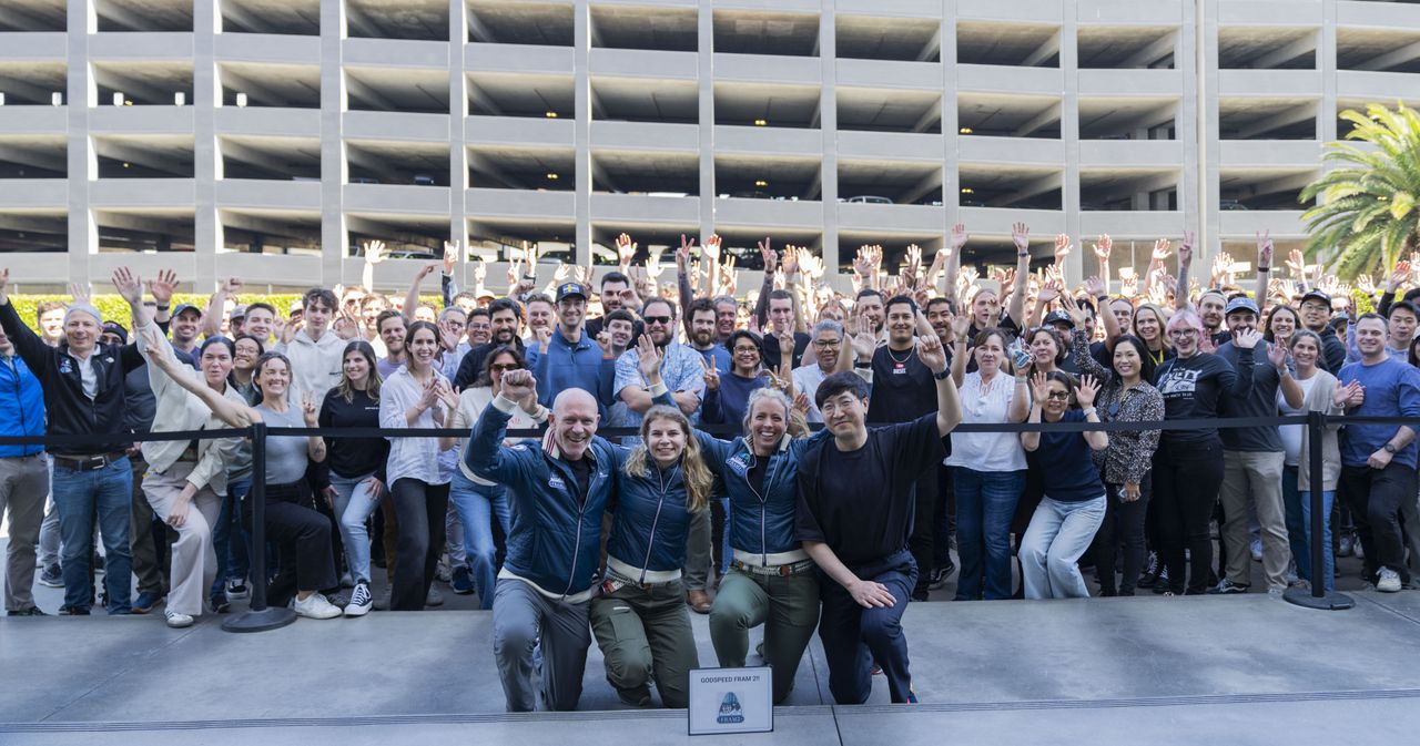 Four humans pose on their knees in front of a crowd of people all raising their hands in excitement.