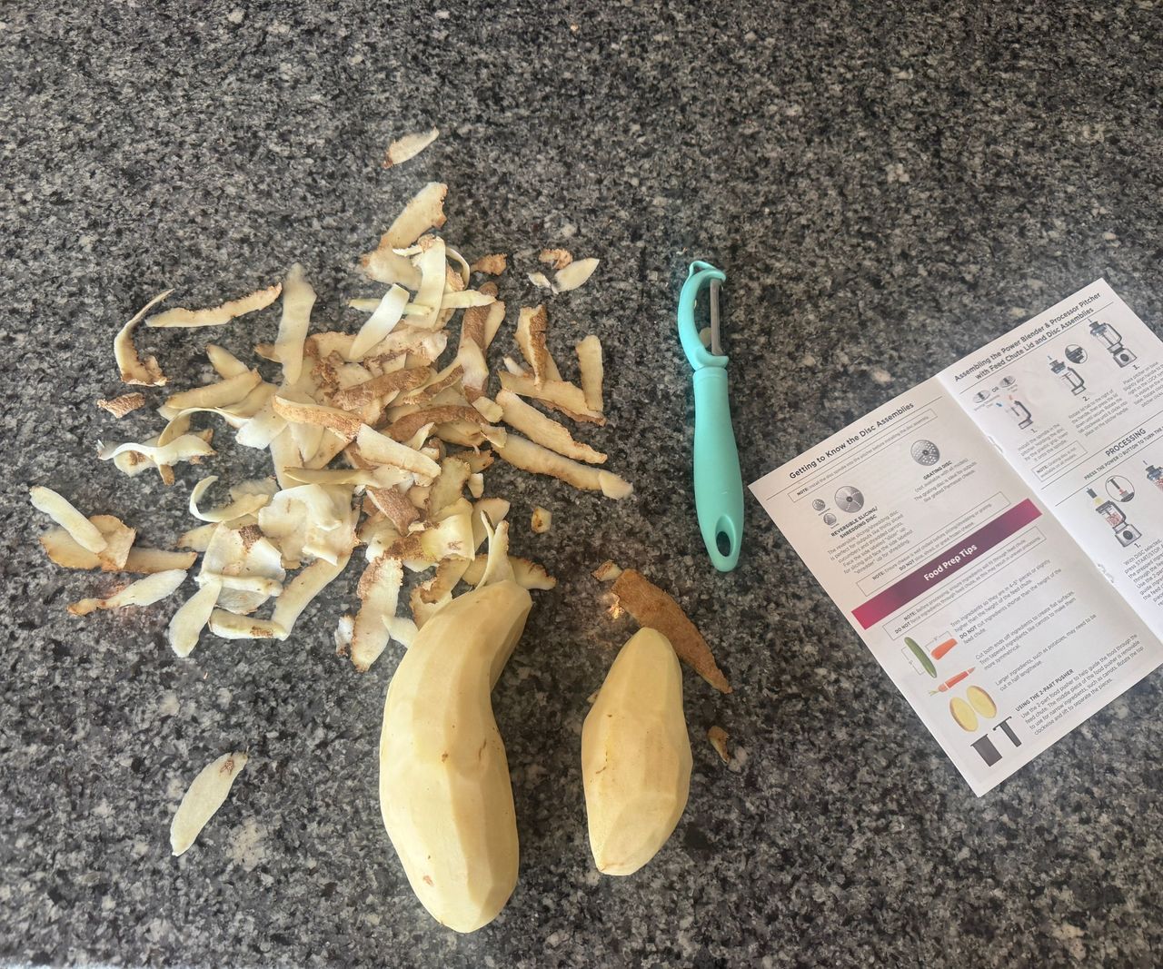 Two potatoes on a kitchen counters, peeled beside their skins, next to a peeler and the open Ninja Foodi Power Blender Ultimate System manual.
